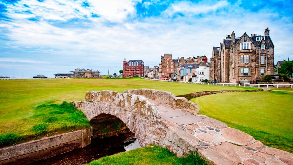 A view of the Swilcan Bridge on the Old Course at St. Andrews, Scotland. The stone bridge crosses a small stream with a green golf course and historic buildings in the background under a partly cloudy sky.