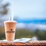 A clear plastic cup filled with a rich, chocolate milkshake sits on a surface. In the background, there are rolling hills leading up to distant, majestic mountains under a clear blue sky.