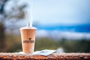 A clear plastic cup filled with a rich, chocolate milkshake sits on a surface. In the background, there are rolling hills leading up to distant, majestic mountains under a clear blue sky.