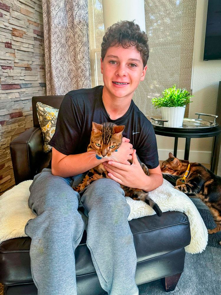 teen holding cat in lap while other cat sniffs him